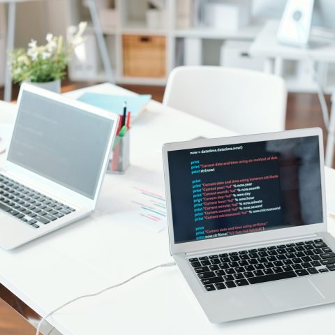 Two laptops on desk with online data on display of one of them inside modern office or classroom with nobody around